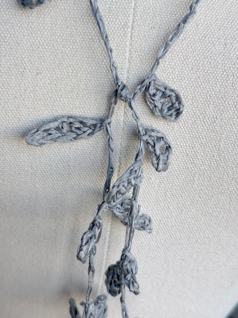 Close-up of a gray crocheted vine or floral garland with leaf-like stitches, laid across a light beige fabric background.
