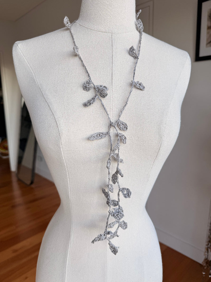 A long silver vine necklace with textured leaf and flower motifs displayed on a white fabric mannequin torso in an indoor room.