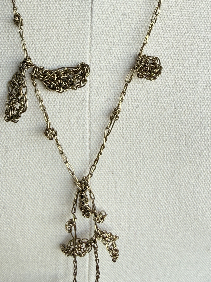 A close-up of a delicate chain necklace with textured, knotted metallic charms in an antique silver or bronze finish, laid flat on a white textured fabric background.